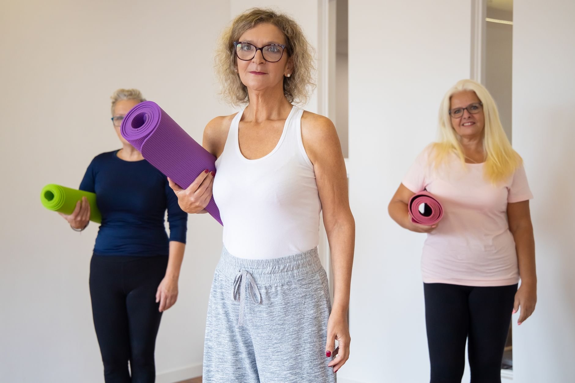 Three senior women holding yoga mats, preparing for a wellness class indoors